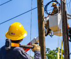 Utility linemen working on electric poles. One lineman is lifted up in the bucket and the other is on the ground with a GEARWRENCH sledgehammer on his shoulder